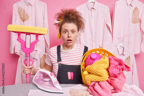 Surprised curly haired woman does domestic work holds mop poses near ironing board with laundry basket shocked to hsve so much work about house dressed in casual clothes. Housekeeping concept photo