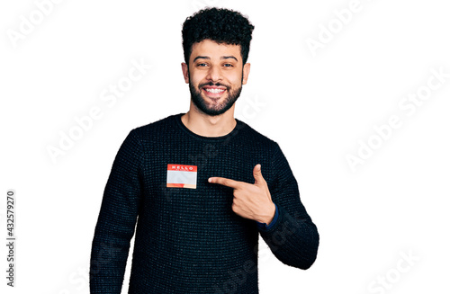 Young arab man with beard wearing hello my name is sticker identification smiling happy pointing with hand and finger photo