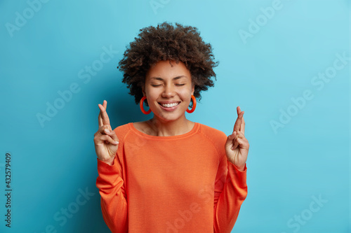 Tablou canvas Lovely cheerful young African American woman with toothy perfect smile keeps fingers crossed believes in good luck wears orange jumper isolated over blue backgound