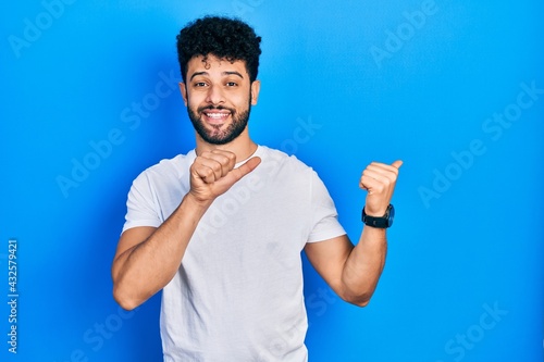 Young arab man with beard wearing casual white t shirt pointing to the back behind with hand and thumbs up, smiling confident