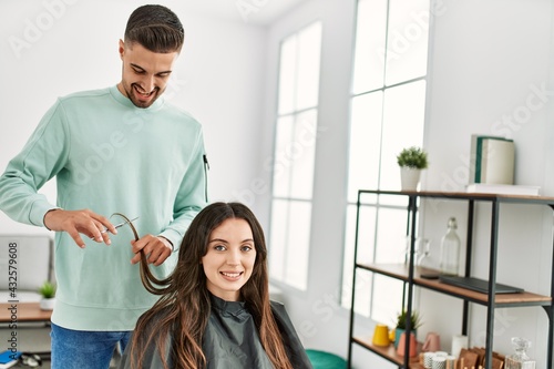 Young hispanic stylist man cutting woman's hair using scissors at beauty center.