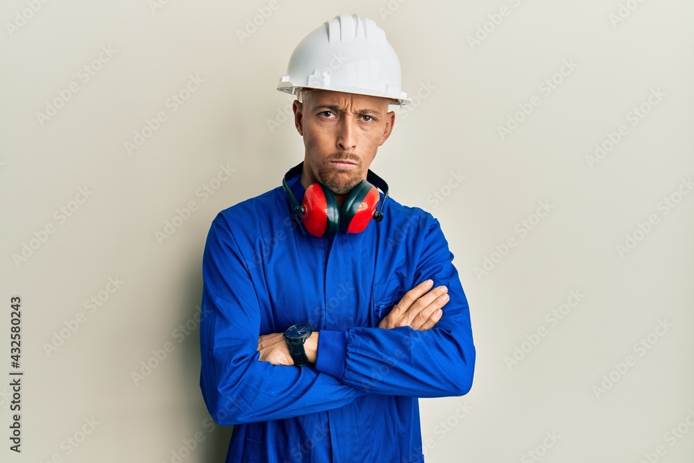 Bald man with beard wearing builder jumpsuit uniform and hardhat skeptic and nervous, disapproving expression on face with crossed arms. negative person.