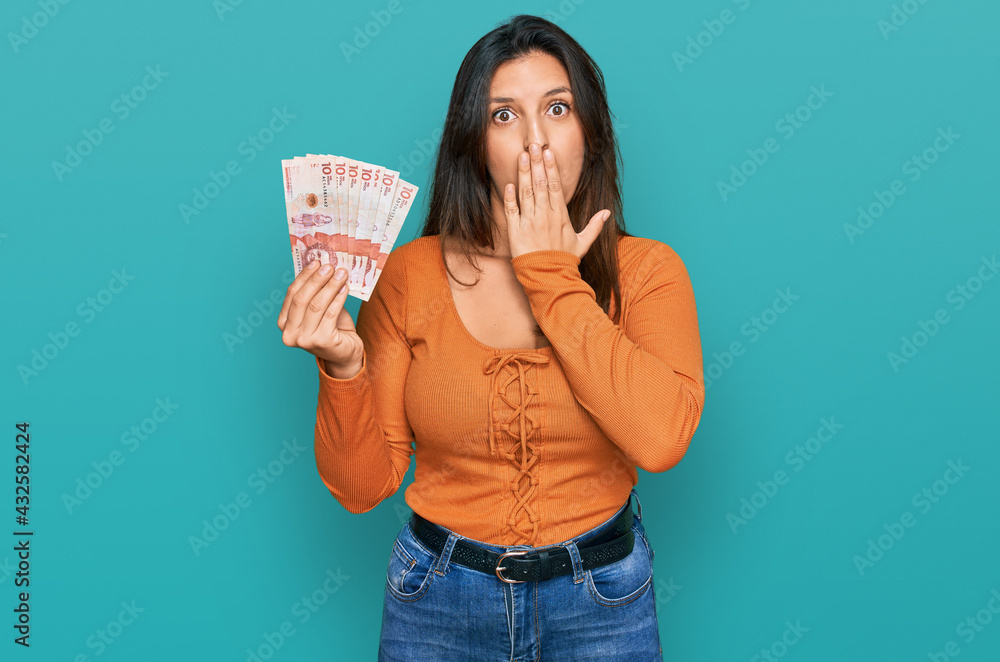 Beautiful hispanic woman holding 10 colombian pesos banknotes covering mouth with hand, shocked and afraid for mistake. surprised expression Beautiful hispanic woman holding 10 colombian pesos banknotes covering mouth with hand, shocked and afraid for mistake. surprised expression
