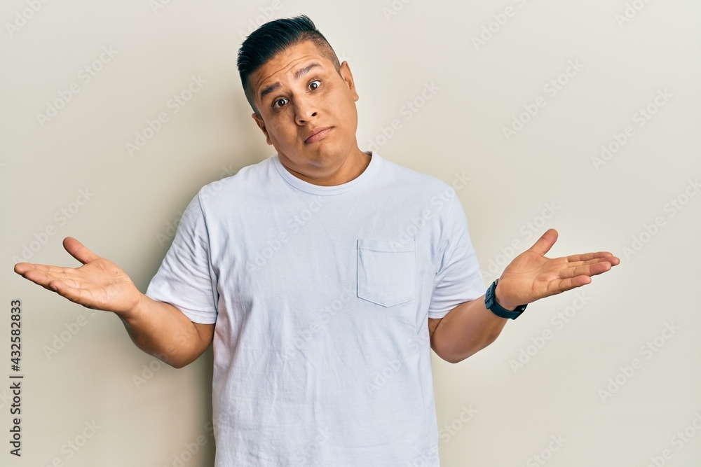 Young latin man wearing casual white t shirt clueless and confused expression with arms and hands raised. doubt concept. Young latin man wearing casual white t shirt clueless and confused expression with arms and hands raised. doubt concept.