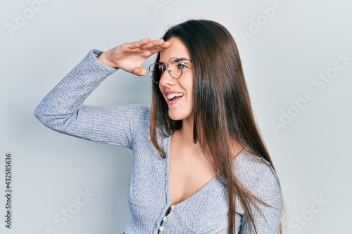 Young brunette teenager wearing casual sweater and glasses very happy and smiling looking far away with hand over head. searching concept.