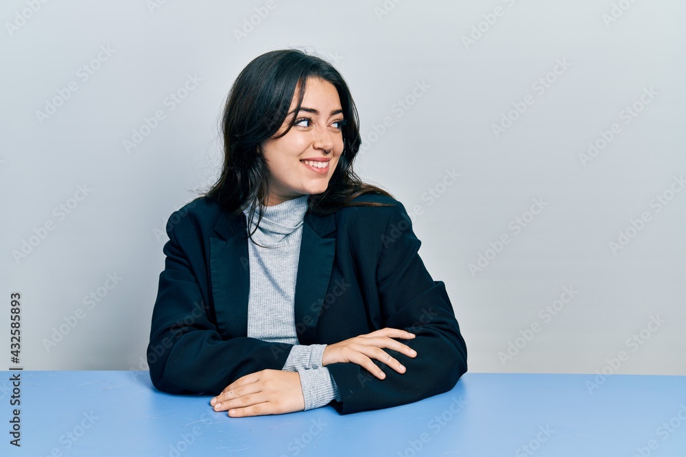 Beautiful hispanic business woman sitting on the table looking away to side with smile on face, natural expression. laughing confident. Beautiful hispanic business woman sitting on the table looking away to side with smile on face, natural expression. laughing confident.