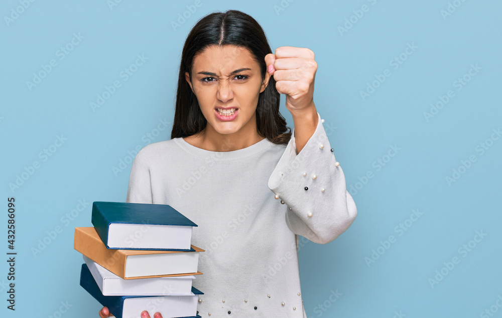 Young hispanic girl holding a pile of books annoyed and frustrated shouting with anger, yelling crazy with anger and hand raised Young hispanic girl holding a pile of books annoyed and frustrated shouting with anger, yelling crazy with anger and hand raised
