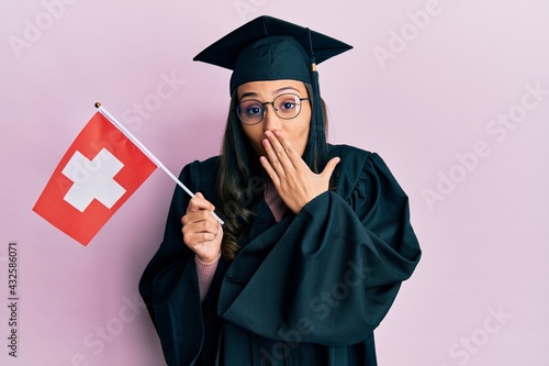 Young hispanic woman wearing graduation uniform holding switzerland flag covering mouth with hand, shocked and afraid for mistake. surprised expression