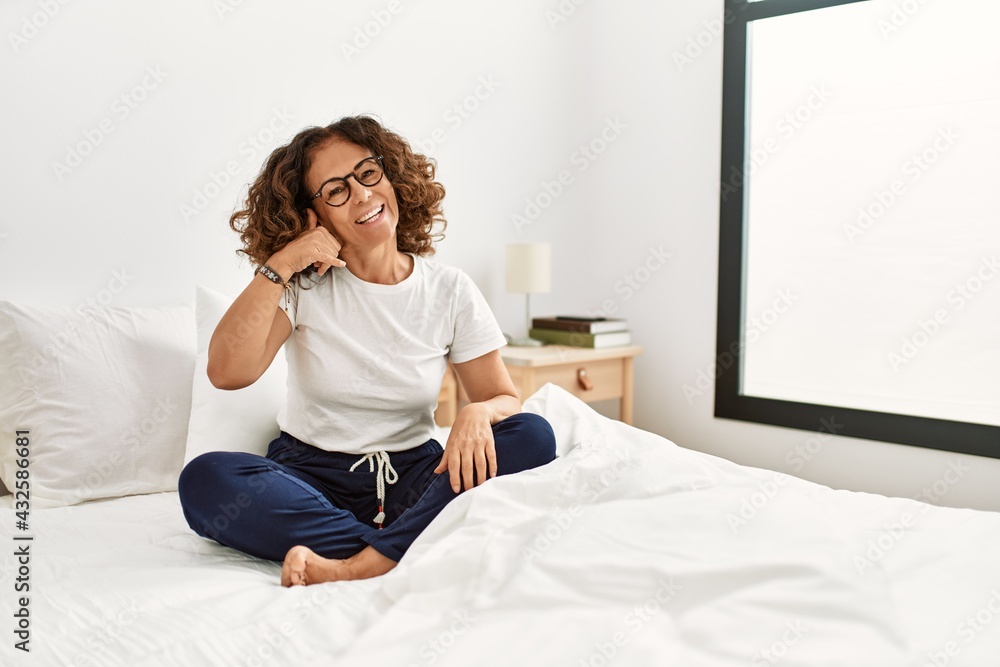 Middle age hispanic woman sitting on the bed at home smiling doing phone gesture with hand and fingers like talking on the telephone. communicating concepts. Middle age hispanic woman sitting on the bed at home smiling doing phone gesture with hand and fingers like talking on the telephone. communicating concepts.