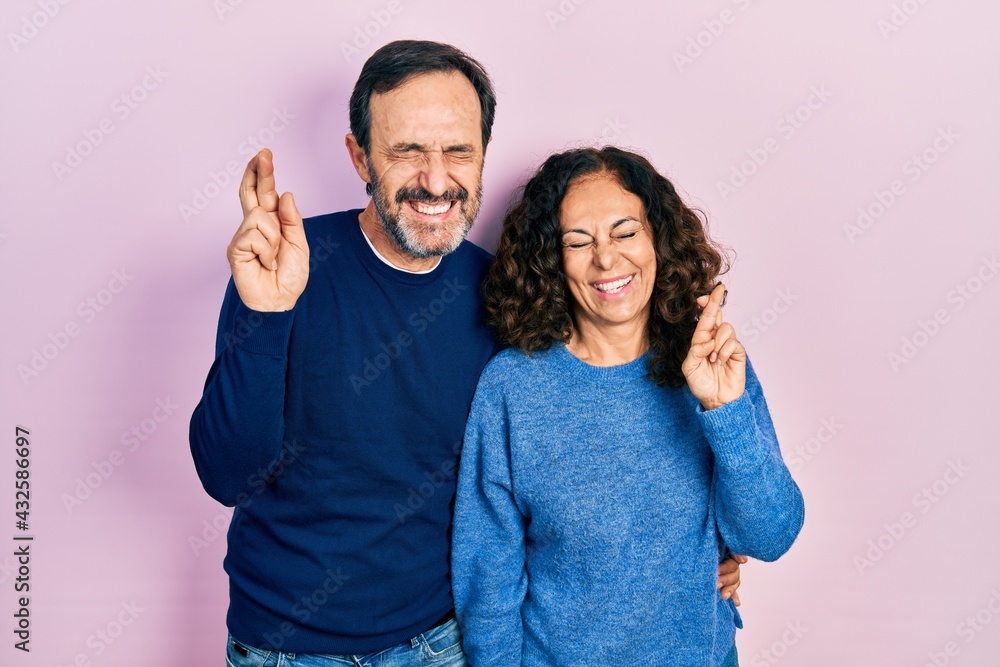 Fototapeta premium Middle age couple of hispanic woman and man hugging and standing together gesturing finger crossed smiling with hope and eyes closed. luck and superstitious concept.