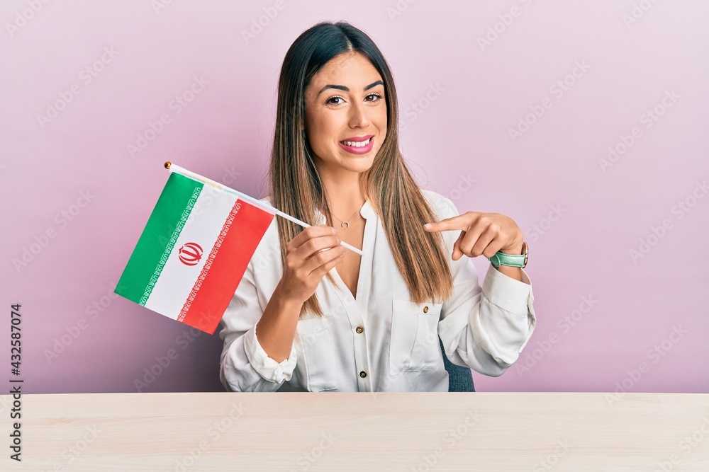 Young hispanic woman holding iran flag sitting on the table smiling happy pointing with hand and finger Young hispanic woman holding iran flag sitting on the table smiling happy pointing with hand and finger