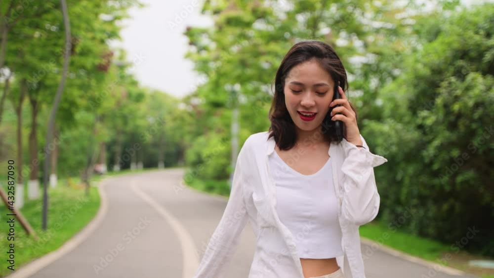 Pretty young asian woman walking on the road in the park outdoor talking on the mobile phone happy people using phone in the nature relax lifestyle Pretty young asian woman walking on the road in the park outdoor talking on the mobile phone happy people using phone in the nature relax lifestyle