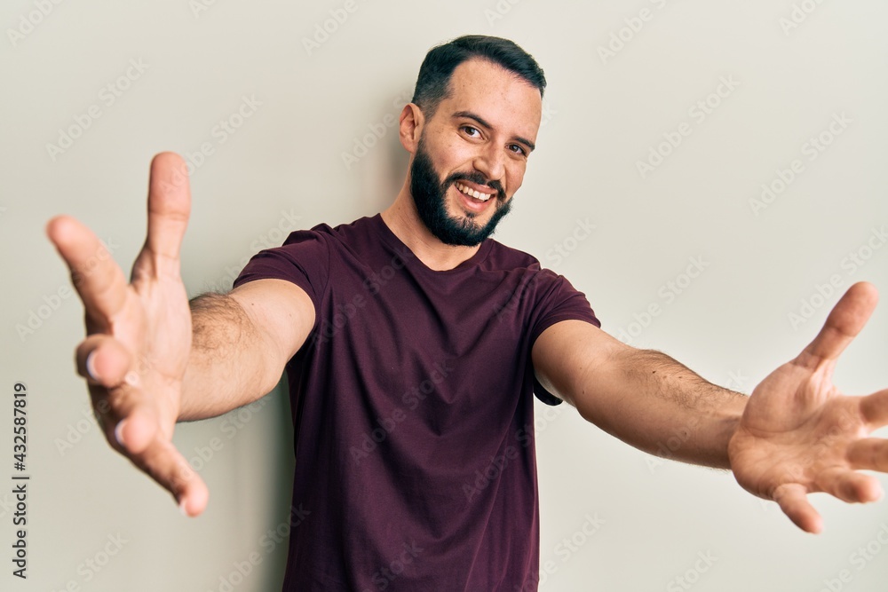 Young man with beard wearing casual t shirt looking at the camera smiling with open arms for hug. cheerful expression embracing happiness. Young man with beard wearing casual t shirt looking at the camera smiling with open arms for hug. cheerful expression embracing happiness.