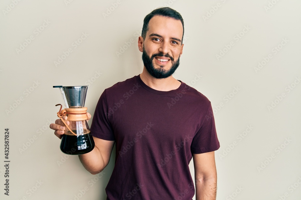 Young man with beard holding coffee maker with filter looking positive and happy standing and smiling with a confident smile showing teeth Young man with beard holding coffee maker with filter looking positive and happy standing and smiling with a confident smile showing teeth