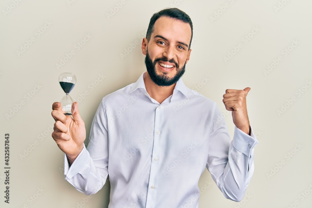 Young man with beard holding sand clock pointing thumb up to the side smiling happy with open mouth Young man with beard holding sand clock pointing thumb up to the side smiling happy with open mouth