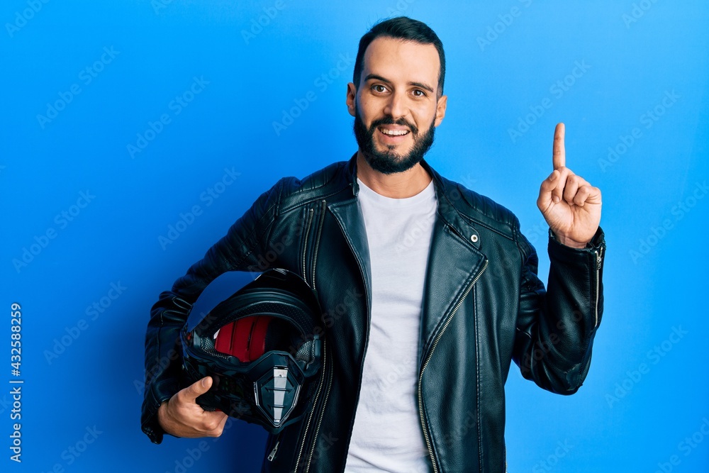 Naklejka premium Young man with beard holding motorcycle helmet surprised with an idea or question pointing finger with happy face, number one