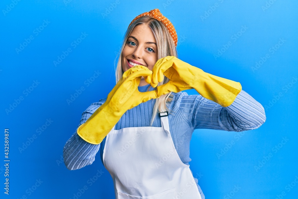 Beautiful blonde woman wearing cleaner apron and gloves smiling in love doing heart symbol shape with hands. romantic concept. Beautiful blonde woman wearing cleaner apron and gloves smiling in love doing heart symbol shape with hands. romantic concept.