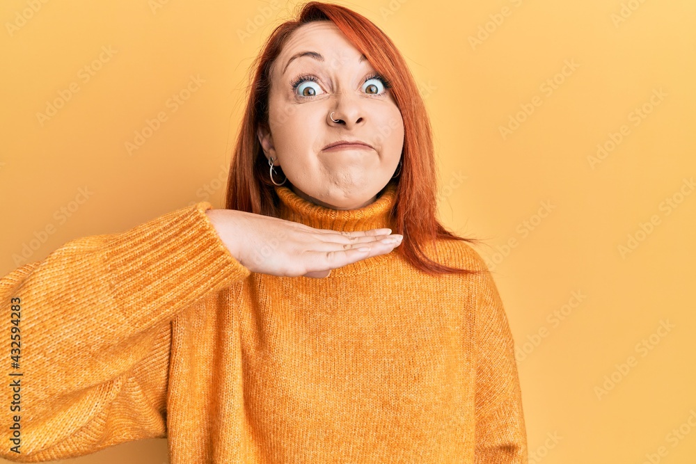 Beautiful redhead woman wearing casual winter sweater over yellow background cutting throat with hand as knife, threaten aggression with furious violence Beautiful redhead woman wearing casual winter sweater over yellow background cutting throat with hand as knife, threaten aggression with furious violence