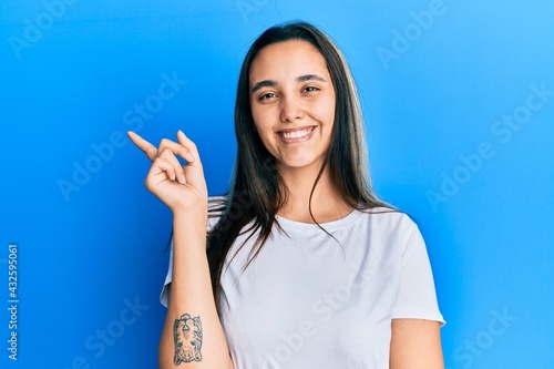 Young hispanic woman wearing casual white t shirt with a big smile on face, pointing with hand and finger to the side looking at the camera.