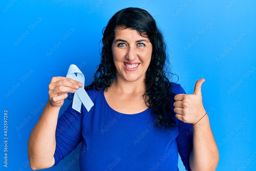 Plus size brunette woman holding blue ribbon smiling happy and positive, thumb up doing excellent and approval sign Plus size brunette woman holding blue ribbon smiling happy and positive, thumb up doing excellent and approval sign