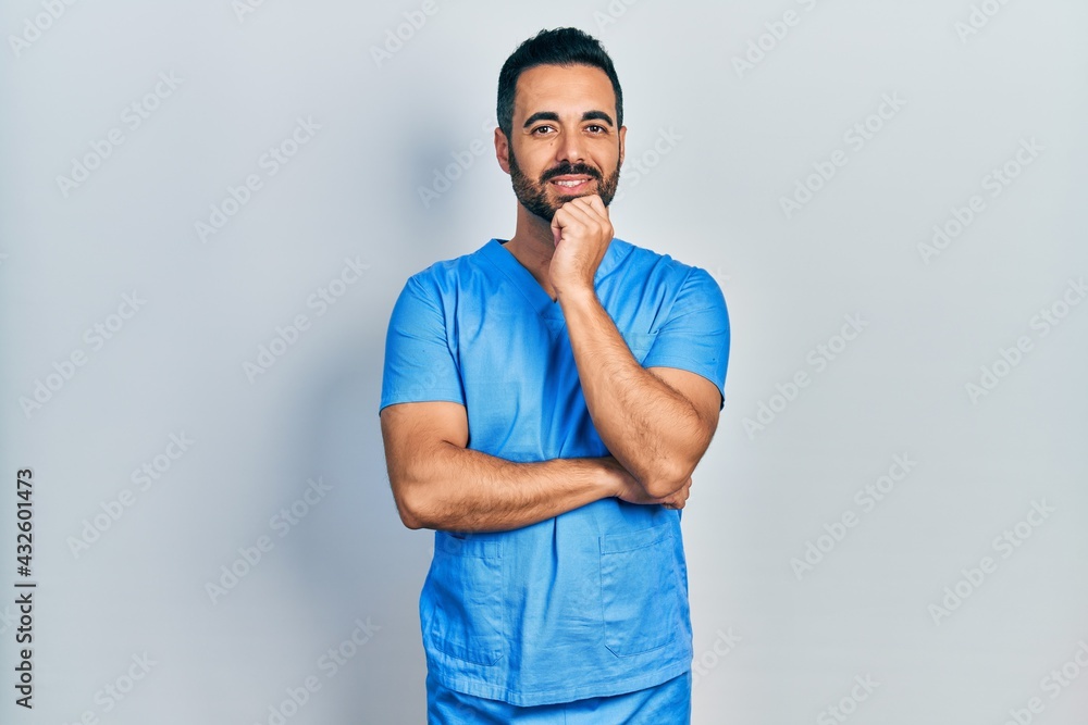 Handsome hispanic man with beard wearing blue male nurse uniform looking confident at the camera with smile with crossed arms and hand raised on chin. thinking positive. Handsome hispanic man with beard wearing blue male nurse uniform looking confident at the camera with smile with crossed arms and hand raised on chin. thinking positive.