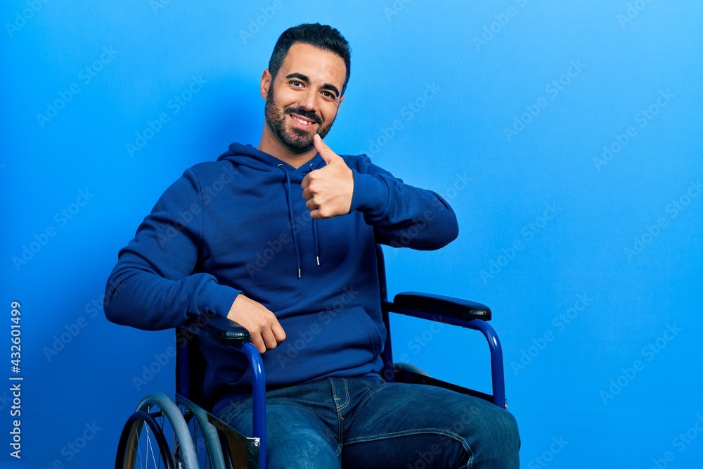 Handsome hispanic man with beard sitting on wheelchair doing happy thumbs up gesture with hand. approving expression looking at the camera showing success.