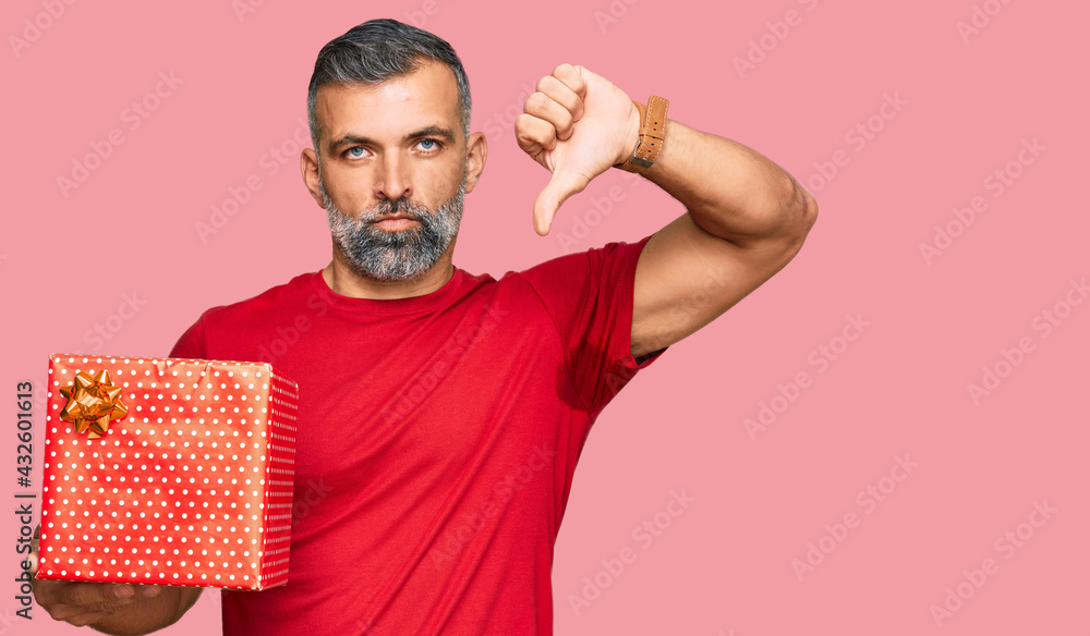 Middle age handsome man holding gift with angry face, negative sign showing dislike with thumbs down, rejection concept Middle age handsome man holding gift with angry face, negative sign showing dislike with thumbs down, rejection concept