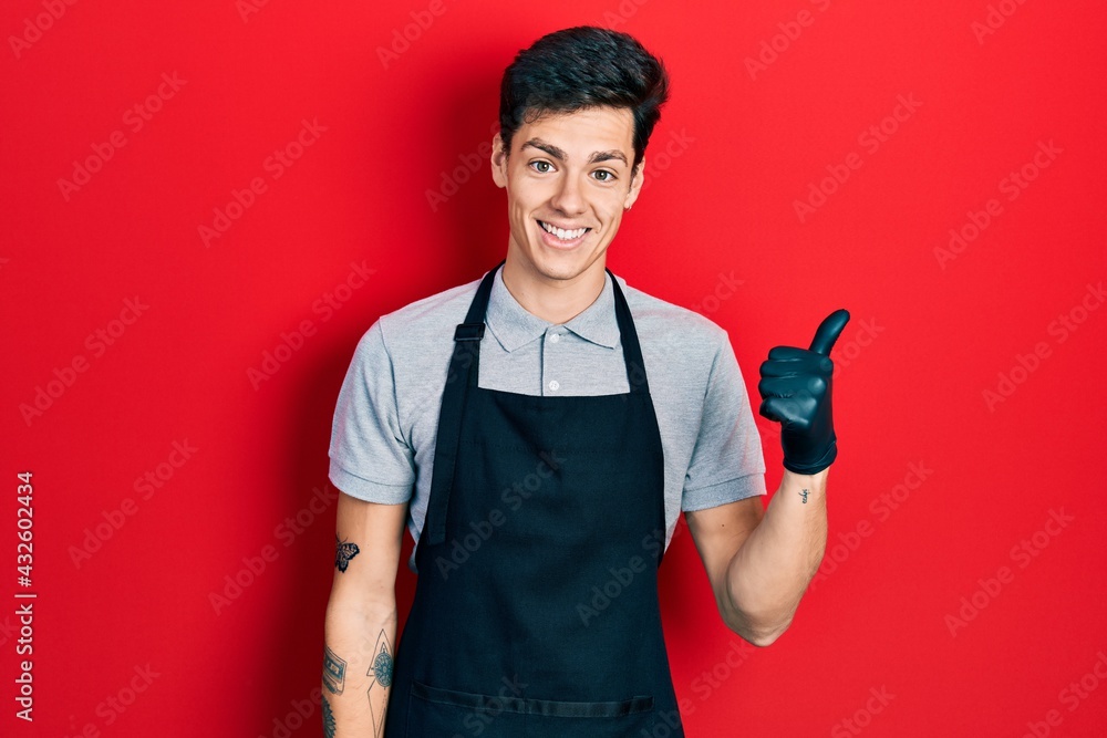Young hispanic man wearing apron smiling with happy face looking and pointing to the side with thumb up. Young hispanic man wearing apron smiling with happy face looking and pointing to the side with thumb up.