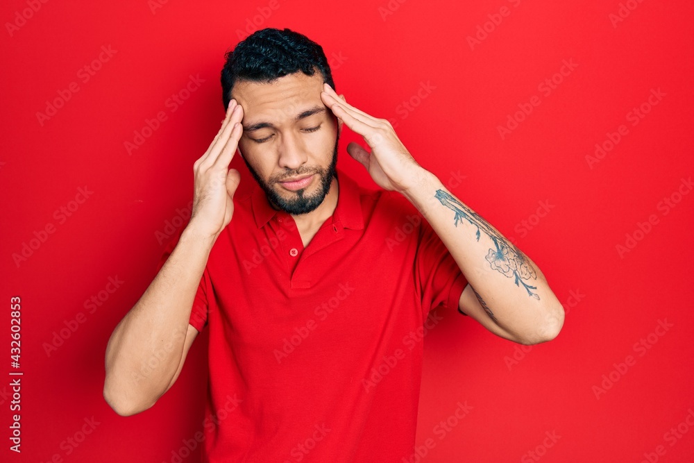 Hispanic man with beard wearing casual red t shirt with hand on head for pain in head because stress. suffering migraine. Hispanic man with beard wearing casual red t shirt with hand on head for pain in head because stress. suffering migraine.