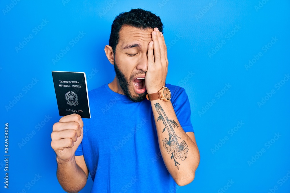 Hispanic man with beard holding italy passport yawning tired covering half face, eye and mouth with hand. face hurts in pain. Hispanic man with beard holding italy passport yawning tired covering half face, eye and mouth with hand. face hurts in pain.