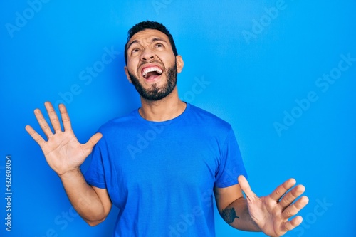 Hispanic man with beard wearing casual blue t shirt crazy and mad shouting and yelling with aggressive expression and arms raised. frustration concept. © Krakenimages.com