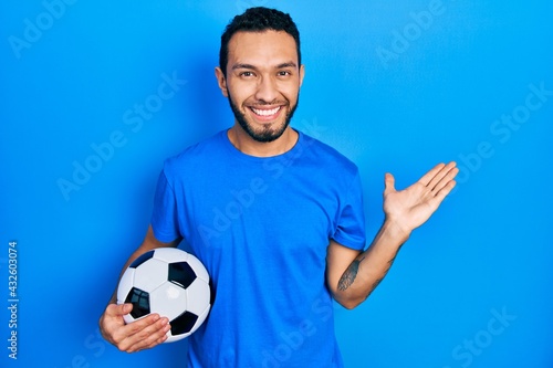 Hispanic man with beard holding soccer ball smiling cheerful presenting and pointing with palm of hand looking at the camera.