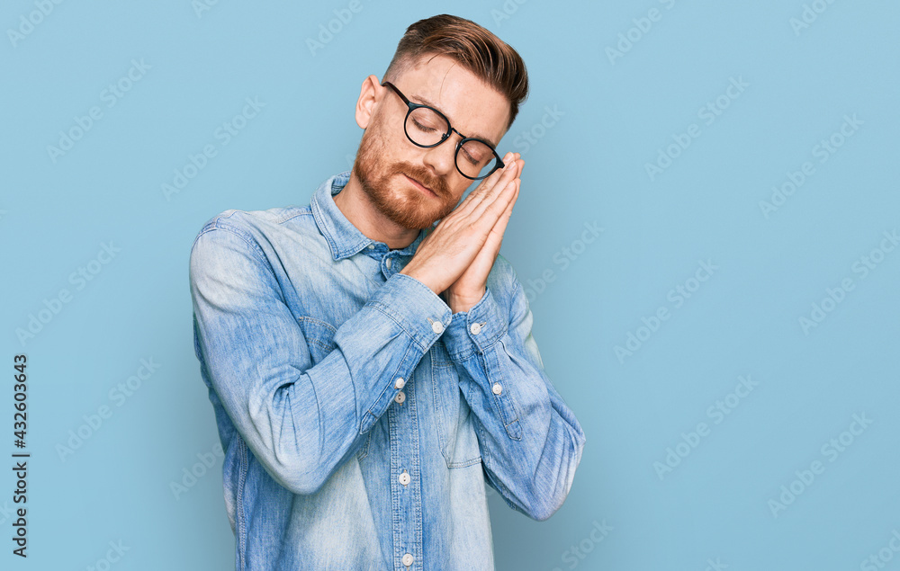 Young redhead man wearing casual denim shirt sleeping tired dreaming and posing with hands together while smiling with closed eyes. Young redhead man wearing casual denim shirt sleeping tired dreaming and posing with hands together while smiling with closed eyes.