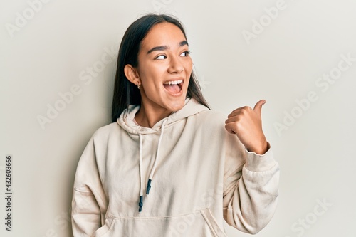Young asian woman wearing casual sweatshirt pointing thumb up to the side smiling happy with open mouth