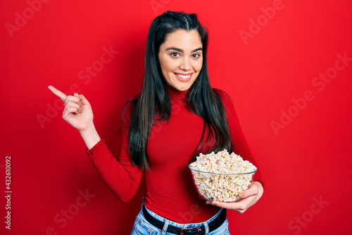 Young hispanic girl eating popcorn smiling happy pointing with hand and finger to the side