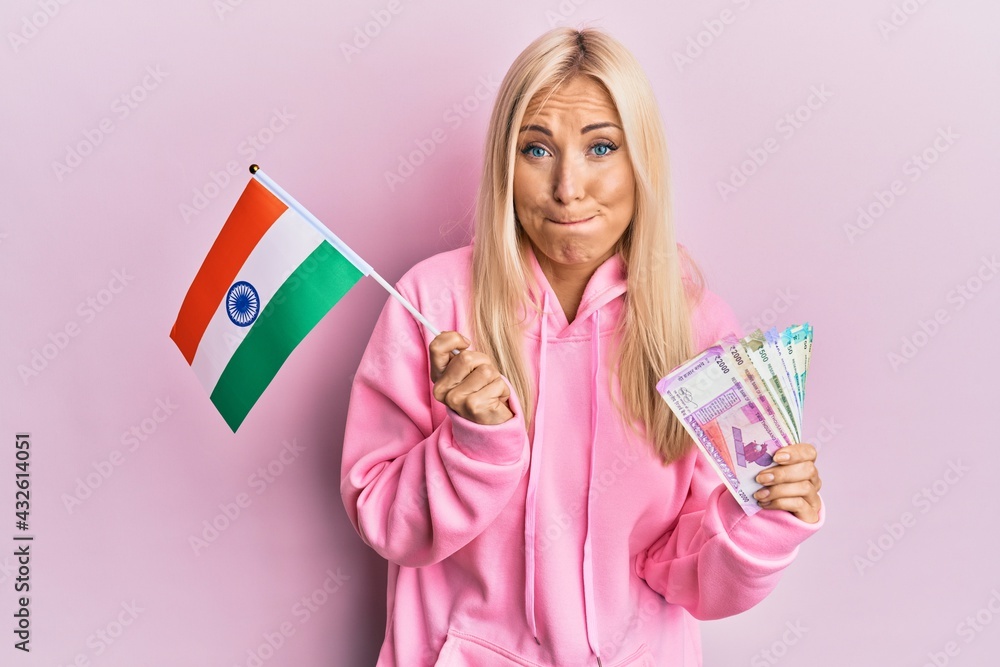 Young blonde woman holding india flag and rupee banknotes puffing cheeks with funny face. mouth inflated with air, catching air. Young blonde woman holding india flag and rupee banknotes puffing cheeks with funny face. mouth inflated with air, catching air.