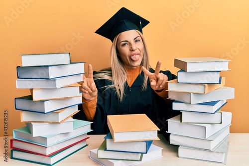 Young caucasian woman wearing graduation ceremony robe sitting on the table smiling with tongue out showing fingers of both hands doing victory sign. number two.