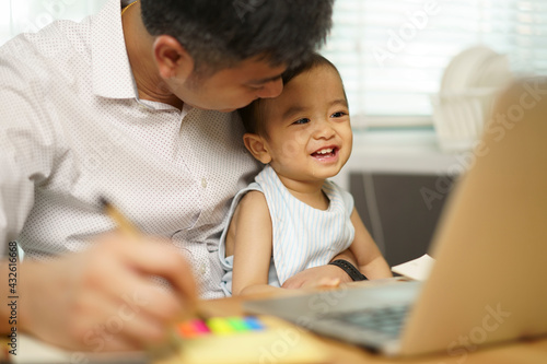 Work from home. Asian young father looking and holding his son on knee. Entrepreneur man working with laptop. A boy sitting with dad at home in kitchen. quarantine on coronavirus pandemic.