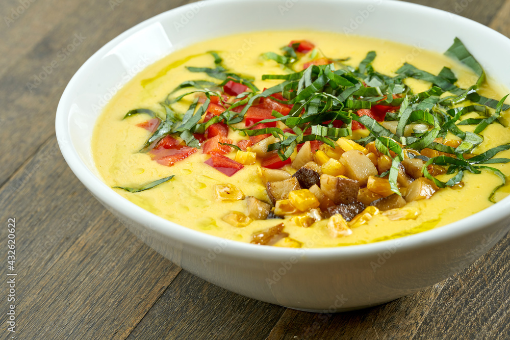 Appetizing and dietary dish - corn soup with cream and fried mushrooms, tomatoes and herbs in a white bowl on a wooden background Appetizing and dietary dish - corn soup with cream and fried mushrooms, tomatoes and herbs in a white bowl on a wooden background