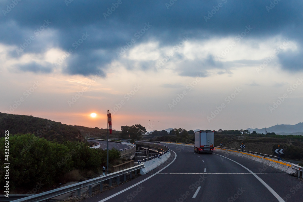 Truck driving on the highway next to a windsock and the sun peeking through the clouds at dawn.