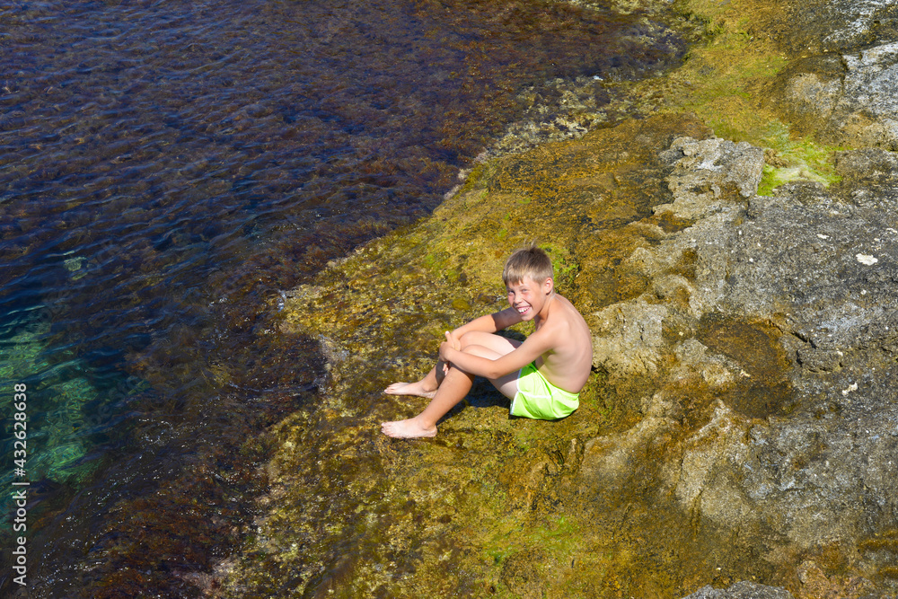 A boy in swimming shorts sits on coral rocks at the water's edge. The smile on the guy's face. Sea shore. Summer sunny day.
A boy in swimming shorts sits on coral rocks at the water's edge. The smile on the guy's face. Sea shore. Summer sunny day.