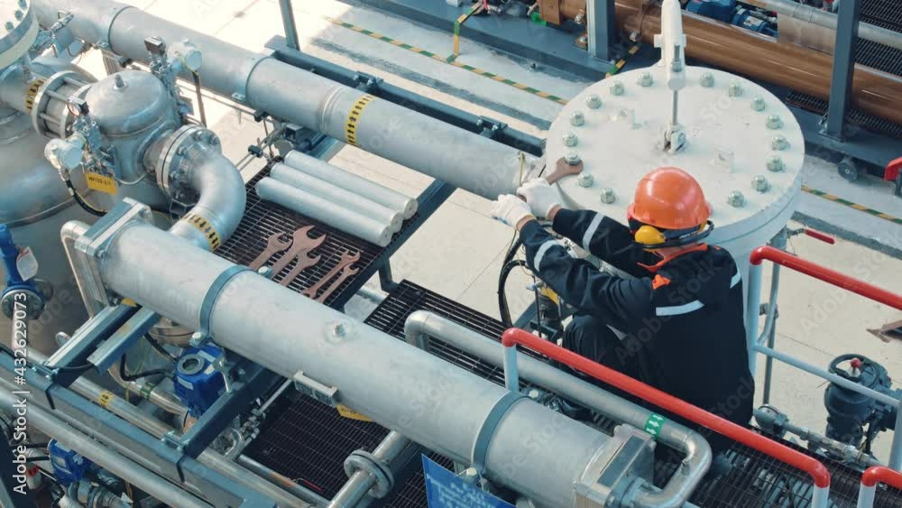 The mechanic is dressed in a black uniform, orange hard hat, goggles and earmuffs. Tightens the nuts with a large wrench on the pressure vessel. Work at an oil refinery. The mechanic is dressed in a black uniform, orange hard hat, goggles and earmuffs. Tightens the nuts with a large wrench on the pressure vessel. Work at an oil refinery.