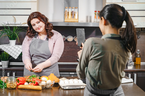 Two girlfriends recording a video or taking a photo on a smartphone for social networks. Plus size female blogger making blog content about diet and healthy food in the kitchen.