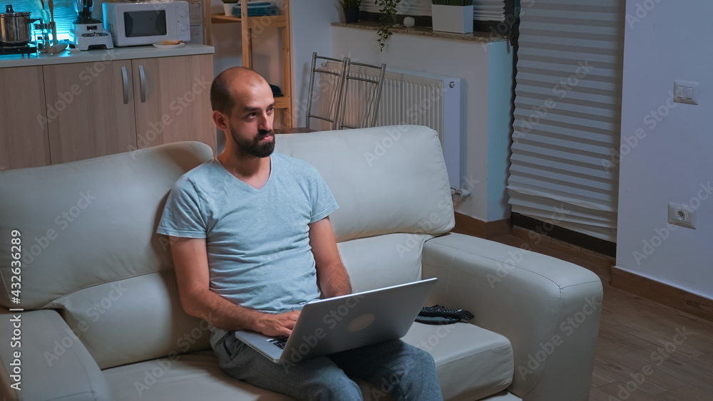 Caucasian male sitting in front of television while working at communication project before deadline on laptop. Tired man browsing on internet using network technology late at night in kitchen Caucasian male sitting in front of television while working at communication project before deadline on laptop. Tired man browsing on internet using network technology late at night in kitchen
