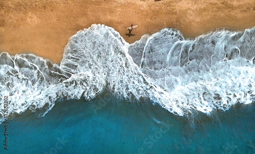 AERIAL, TOP DOWN: Unrecognizable surfer walking along the exotic sandy beach.
