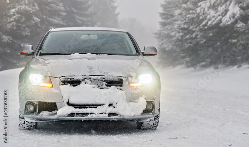 Silver car parked on snow covered winter road  blurred trees background  front view  headlights shining