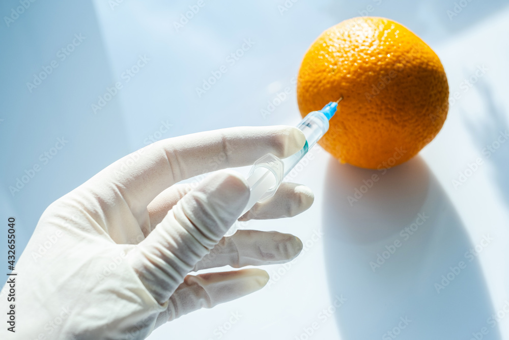 laboratory assistant injects chemicals with a syringe into an orange. fruits are processed for long-term storage. processed edible vegetables and fruits on a white background. genetic engineering layo laboratory assistant injects chemicals with a syringe into an orange. fruits are processed for long-term storage. processed edible vegetables and fruits on a white background. genetic engineering layo