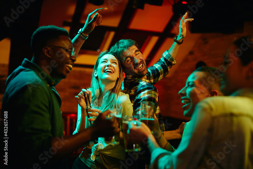Group of cheerful friends drinking and dancing while gathering in pub at night.