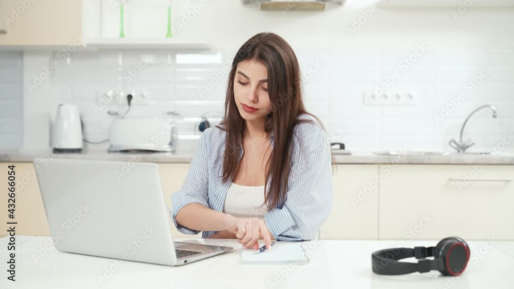 Front view of attentive and beautiful young brunette woman sitting and working on laptop at home at writing some information on notebook. Concept of working or studying at home during quarantine. Front view of attentive and beautiful young brunette woman sitting and working on laptop at home at writing some information on notebook. Concept of working or studying at home during quarantine.