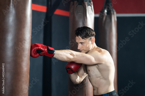 Shirtless handsome Caucasian men boxer with strong muscle training boxing punch in side of the gym with sandbag. Cardio sport workout activity for good health and build up body and physical strength.